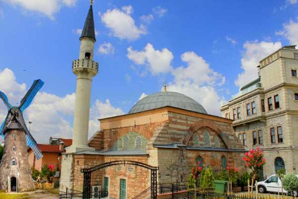 Osmanlı camisi ve yanında yel değirmeni var. Rüya açıklamasına istinaden. Yapay bir resim. Sağ tarafta üç katlı bir bina ve binanın önünde beyaz bir otomobil vard.